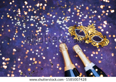 Two champagne bottles, golden carnival mask and confetti stars on purple-blue backround. Flat lay of Christmas, New Year, Purim, Carnival celebration concept.