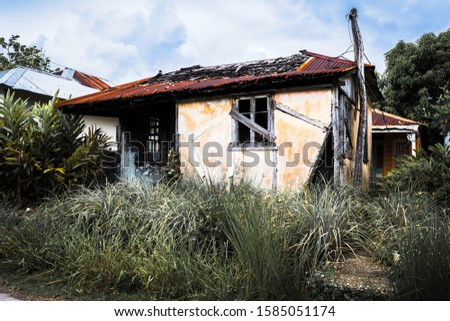 Abandoned rusty old vintage countryside house. Burned concrete and wooden home structure with zinc roof/ roofing in rural residential lot with overgrown grass plants. Damaged small country dwelling. Royalty-Free Stock Photo #1585051174