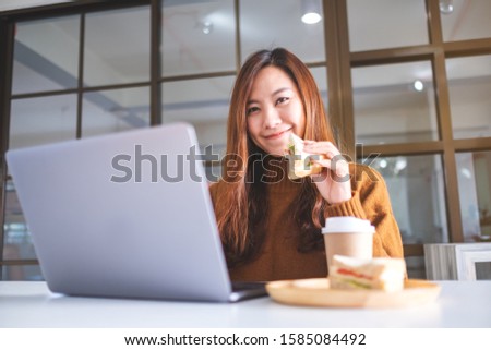 An asian woman holding and eating whole wheat sandwich while working on laptop computer