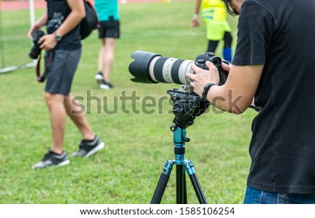 Unrecognised photographer man with zoom lens, tripod, and professional digital camera. He takes sports action photos of soccer player playing football.
