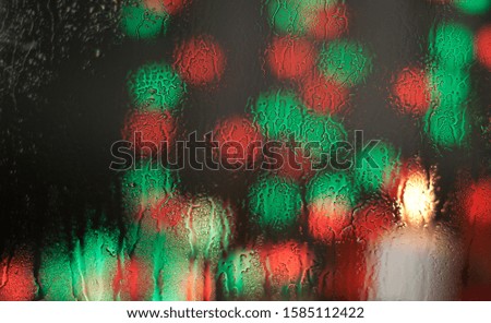 On a transparent glass, drops of water flow from the rain from the street. Through it, blurry bokeh and multi-colored spots from the New Year's garland are visible