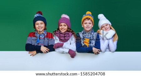 Four beautiful kids in winter clothes posing together with white board at studio. Green background. Winter fashion, winter activities. Christmas and New Year. Copy space.