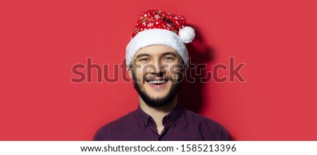 Portrait of young smiling man wearing Christmas hat with snowflakes isolated on red background.