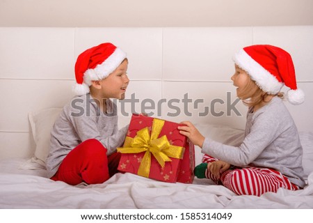 Funny boy and girl having fun in bedroom on Christmas morning. Kids in Santa Clause red hats open gift box. Happy xmas and New Year. Children playing in bed.