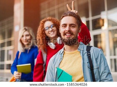 Happy young man in casual outfit smiling and looking at camera while female friend making horn gesture on sunny day outside university