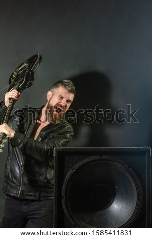 Guitarist breaks guitar at music column. Angry man with beard breaks guitar at music speakers. Emotions, aggression, musical instruments. Guitar player at rock concert. Musician with electric guitar.