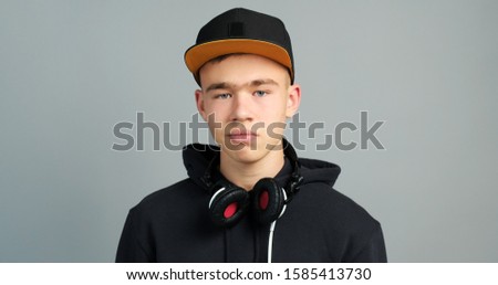 Portrait of teenager boy in cap with headphones smiling looks and the camera at the interlocutor on a grey background. Modern teens lifestyle. Copy space. 