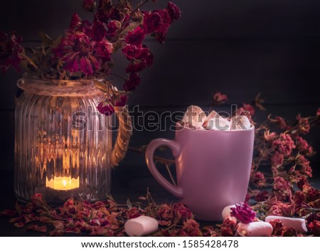 Dark photo. Hot drink with marshmallows on a dark background among a burning candle and red rosebuds. Cozy evening atmosphere.