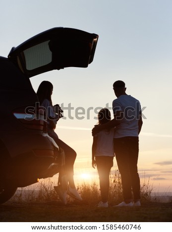 Silhouette of the family of three enjoying the sunset on the top of hill outside the city, woman is sitting in the car trunk, holding jack russell terrier, man is standing embracing his daughter