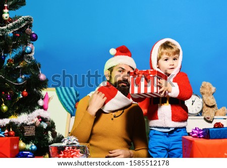 Santa and little assistant among gift boxes near Christmas tree. Christmas eve and surprised concept. Christmas family opens presents on blue background. Man with beard and curious face plays with son