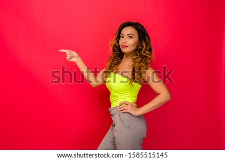 Pleased brunette woman green top pointing finger on copyspace while looking at the camera over red background. - image