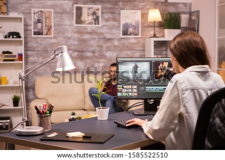 Creative female videographer working on a movie using her home computer. Boyfriend relaxing on sofa in the background.