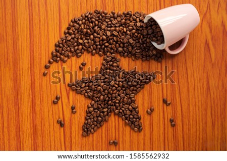 Spilled coffee beans from the pink ceramic mug forming a star shape on a wooden table. Background concept for Christmas greeting card, winter morning, coffee addiction.