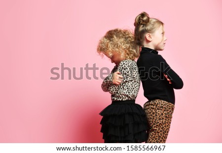 Two offended children kids girls best friends sisters in leopard print clothes pants and sweater are standing with their backs to each other on pink background with copy space