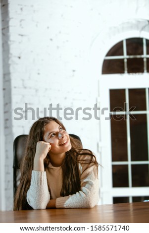 Portrait of young caucasian girl in casual clothes looks dreamful, cute and happy. Looking up and thinking, sitting indoors at the wooden table. Concept of future, target, dreams, visualisation.