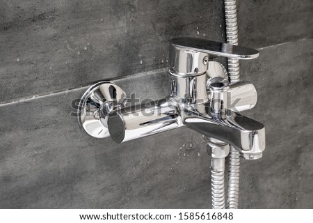 Chrome metal faucet for hot and cold water, with a shower hose, mounted in a wall covered with gray tile imitating concrete with water drops in a modern bathroom