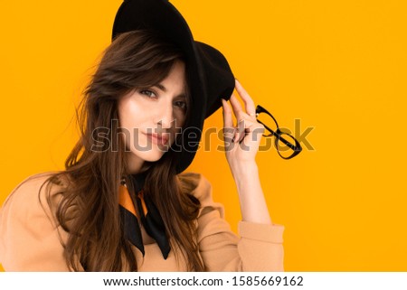 attractive brunette young woman in a black hat and blouse and with glasses on an orange background