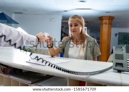 Stock photo of a young blonde girl picking up her room card at a hotel reception while the receptionist is on the phone