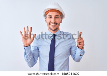 Young business man wearing contractor safety helmet over isolated background showing and pointing up with fingers number six while smiling confident and happy.
