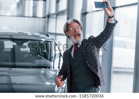 Positive senior man with grey hair taking selfie against modern car indoors.