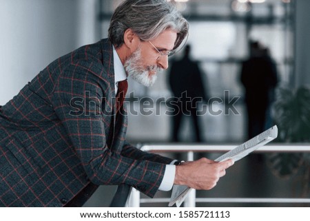 Senior businessman in suit and tie with gray hair and beard holds newspaper in hands.