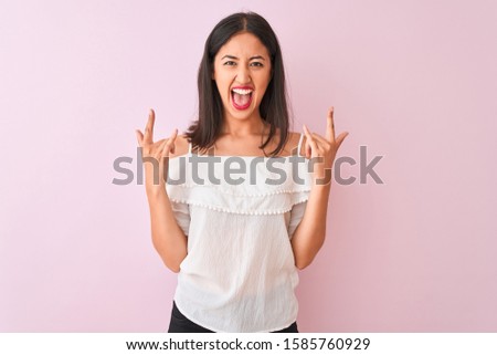 Beautiful chinese woman wearing white t-shirt standing over isolated pink background shouting with crazy expression doing rock symbol with hands up. Music star. Heavy concept.
