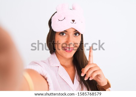 Girl wearing pajama and sleep mask make selfie by camera over isolated white background surprised with an idea or question pointing finger with happy face, number one