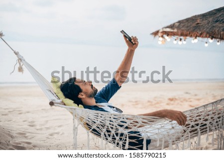 A man enjoys calm, lies in a hammock with a phone on the background of the ocean and sunset. A handsome guy looks, reads on the phone at sunset on a tropical island beach.