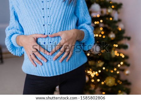 Close up of pregnant woman making heart shape gesture with hands on her belly at home over christmas tree background.