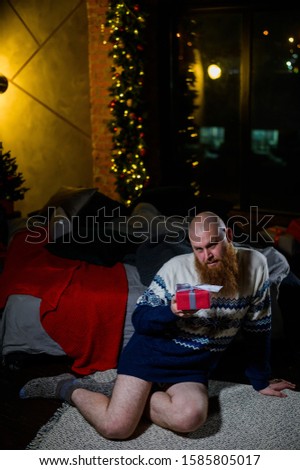 A man with a long red beard sits on the floor without pants in a winter sweater. man is holding a wrapped present on the background of New Year decorations and lights. Christmas tree. Parody, humor.