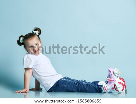 Happy cheerful frolic kid girl with funny buns and in t-shirt, blue jeans and roller skates on her feet is sitting sideways to us looking at us and smiling. Copy space