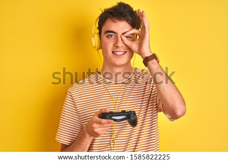 Teenager boy playing video games using gamepad over isolated yellow background with happy face smiling doing ok sign with hand on eye looking through fingers