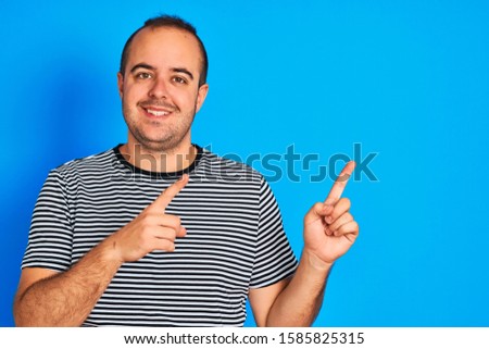 Young man wearing striped navy t-shirt standing over isolated blue background smiling and looking at the camera pointing with two hands and fingers to the side.