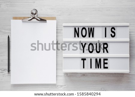 'Now is your time' words on a modern board, clipboard with blank sheet of paper on a white wooden background, top view. Overhead, from above, flat lay. Copy space.