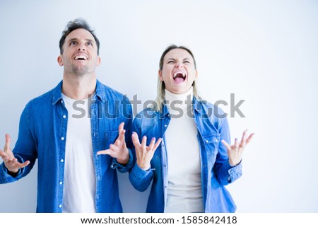 Young beautiful couple wearing denim shirt standing over isolated white background crazy and mad shouting and yelling with aggressive expression and arms raised. Frustration concept.