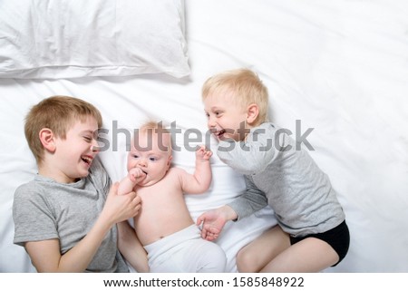 Two older brothers play and laugh with the youngest baby in a white bed. Happy childhood, big family