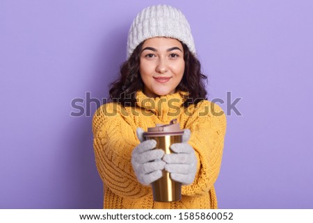 Portrait of pleasant attractive magnetic woman looking directly at camera, holding thermo pot in both hands, warming her hands, wearing white gloves hat and yellow sweater. Winter holidays concept.