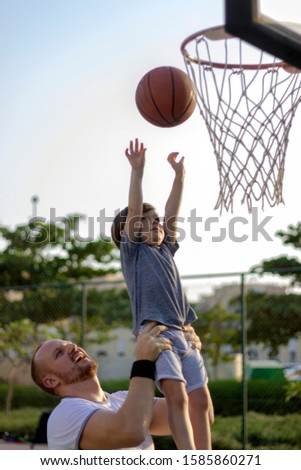 Dad and son playing basketball together.Father raised his son up in the air.Low angle image of son in his father's arms throwing a basketball into the hoop.Shot of happy family bonding.Copy space.