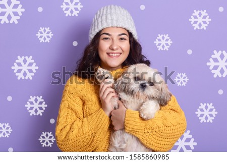 Horizontal indoor picture of cheerful smiling curly haired woman standing isolated over lilac background, holding Maltese dog in hands, having peaceful facial expression. People and animals concept.