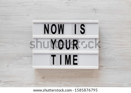'Now is your time' words on a lightbox on a white wooden surface, top view. Overhead, from above, flat lay. 