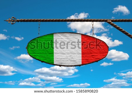 Italy flag on a signboard. Oval signboard colors Italy flag hanging on a metal forged structure. Template on a background of blue sky with clouds. Blank for creativity and design.