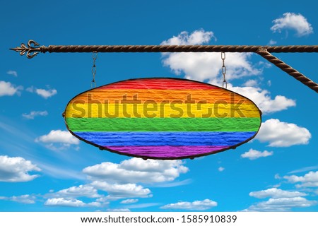 LGBT flag on a signboard. Oval signboard colors LGBT flag hanging on a metal forged structure. Template on a background of blue sky with clouds. Blank for creativity and design.