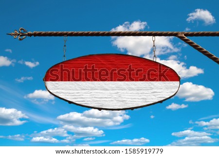 Monaco flag on a signboard. Oval signboard colors Monaco flag hanging on a metal forged structure. Template on a background of blue sky with clouds. Blank for creativity and design.