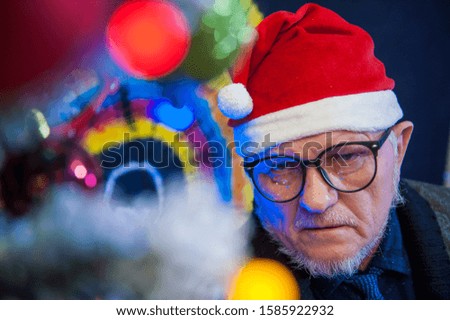 elderly man in glasses with gray hair and beard next to luminous and blinking Christmas lights. Glasses reflect flashing garlands with lights. Christmas tree, colored background, Santa Claus hat