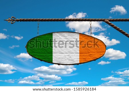 Ireland flag on a signboard. Oval signboard colors Ireland flag hanging on a metal forged structure. Template on a background of blue sky with clouds. Blank for creativity and design.