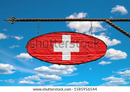 Switzerland flag on signboard. Oval signboard colors flag Switzerland hangs on a metal forged structure. Template on a background of blue sky with clouds. Blank for creativity and design.