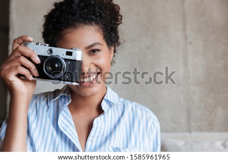 Smiling attractive young african woman wearing shirt sitting on a couch at home, holding photo camera