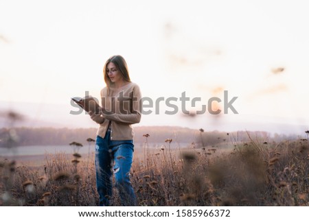 Beautiful brunette girl in glasses dressed in casual clotehs walks among the meadow and reading a book, colorful forest on the background, autumn time