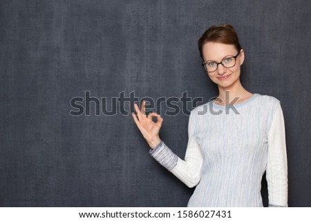 Studio half-length portrait of happy satisfied girl with glasses, smiling cheerfully, showing ok gesture or counting something and showing zero with fingers, over gray background, copy space on left