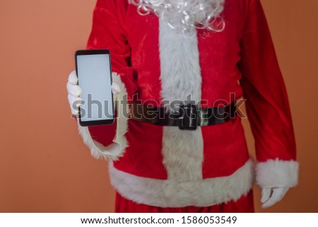 Closeup on a busy Father Santa Claus using showing mobile smart phone and holding shopping paper bag. Mockup copy space modern design display festive decoration background photography. Happy New Year!